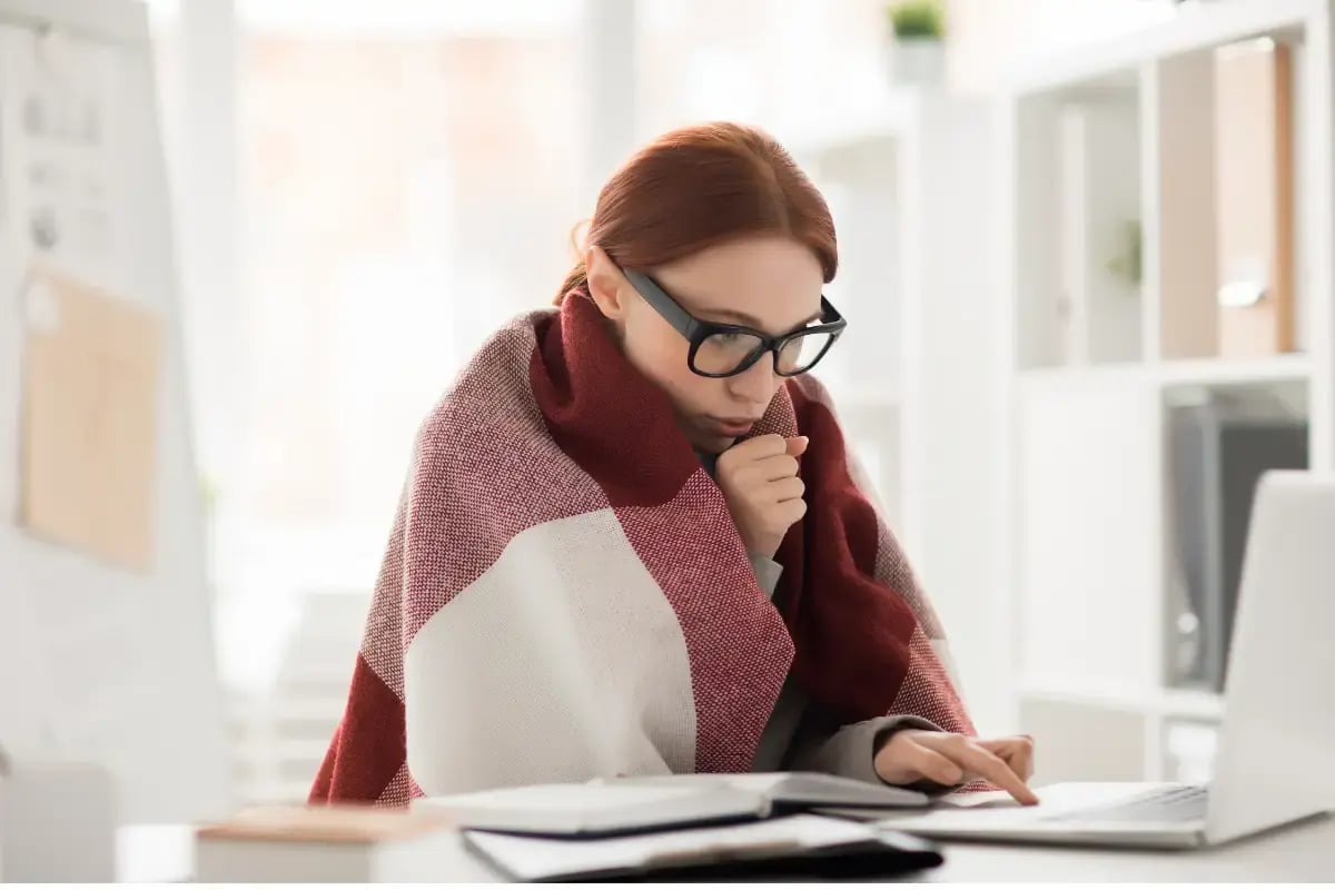 A female employee types on her laptop in a chilly office environment, highlighting why employers benefit from having a space heaters in the workplace policy.